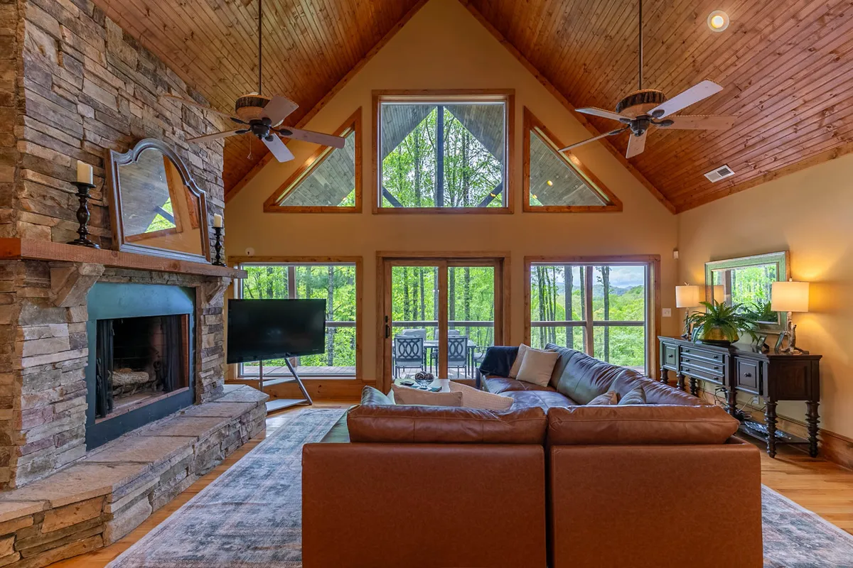 A spacious living room featuring a prominent stone fireplace, large windows showcasing mountain views, a plush leather couch, and a warm wooden ceiling. The natural light and cozy furnishings make it a perfect gathering spot for relaxation and storytelling.