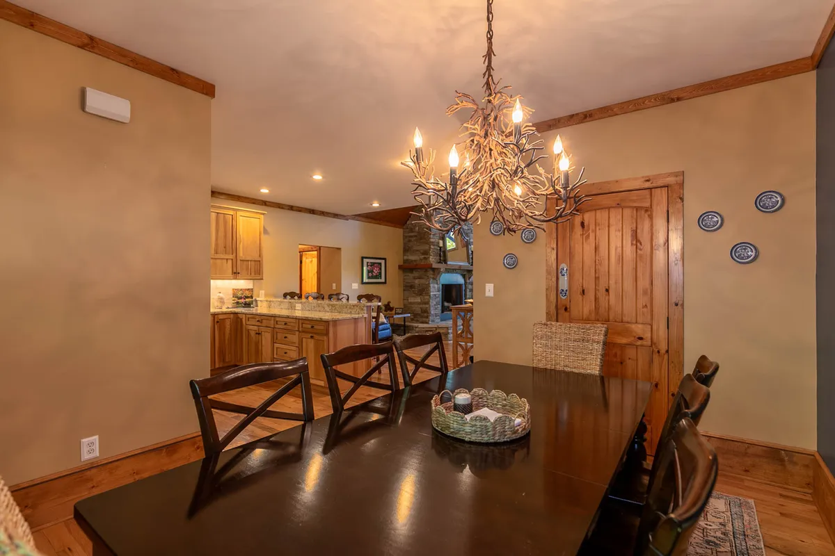 A warmly lit dining area featuring a wooden table set beneath a glowing chandelier, with views into the adjacent kitchen. The layout invites large family dinners, celebrations, and lingering conversations.