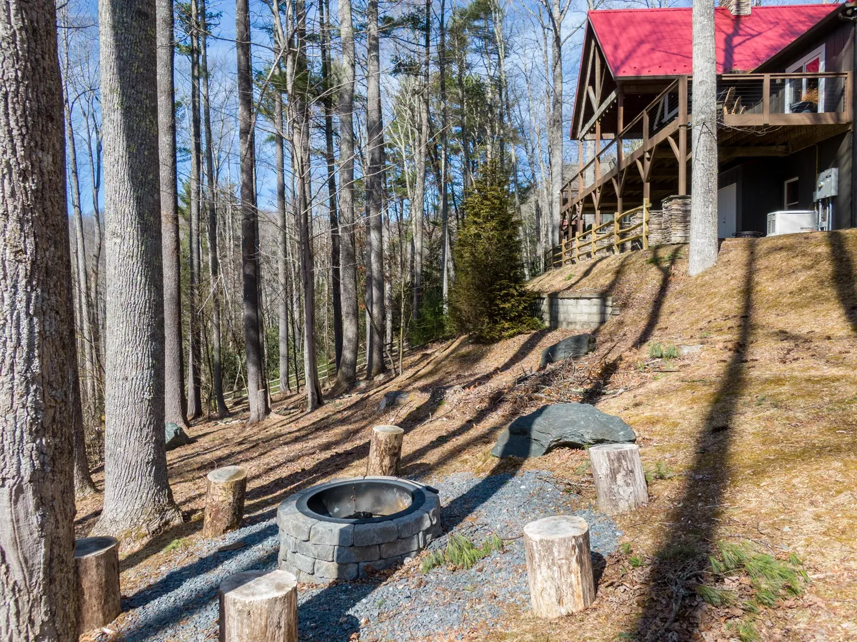 A cozy fire pit area is set in a wooded outdoor space, with stump seating and a view of the cabin's red roof. The scene invites campfire gatherings, storytelling, and stargazing, all quintessential family vacation experiences.