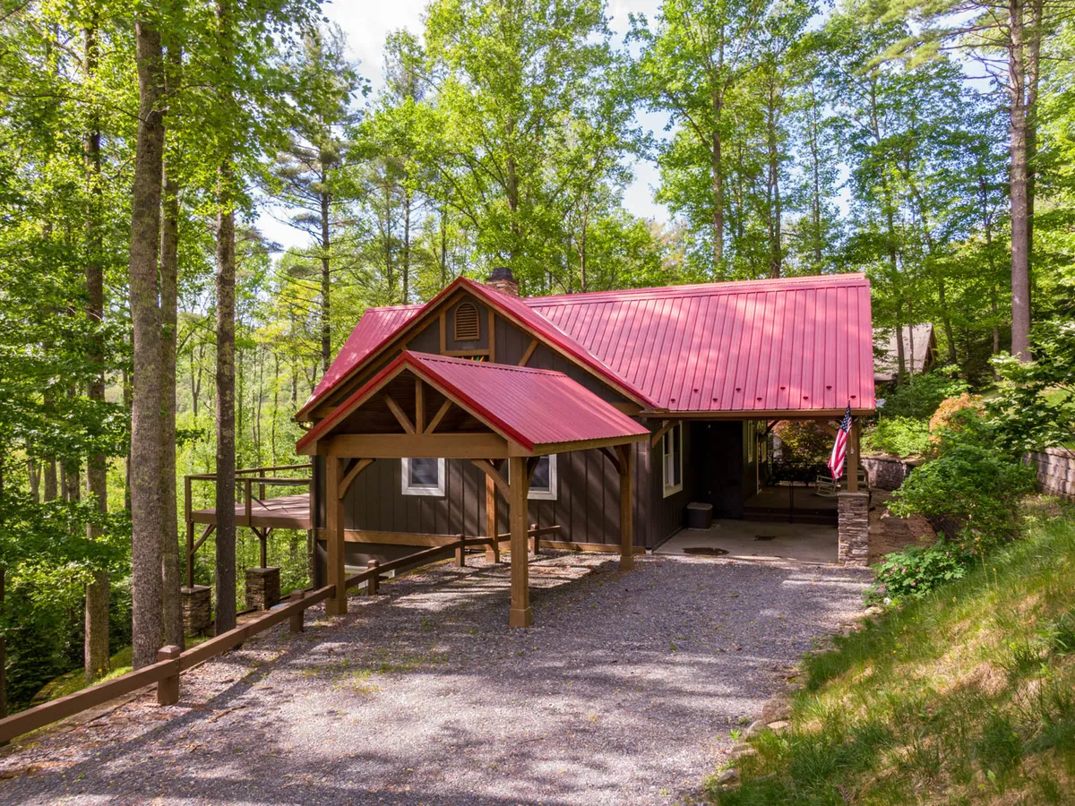 This image showcases the home's exterior, featuring a red metal roof, a wooden porch, stone accents, and the surrounding trees. The setting is inviting and cozy, with open spaces that hint at a large yard where children and pets can play. The porch suggests a great spot for relaxing after a day of local exploration.