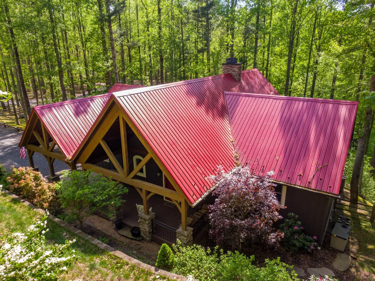 A charming exterior view featuring a red metal roof, wooden beams, stone accents, and lush green trees. The inviting porch and natural surroundings evoke a sense of arrival and community gatherings.