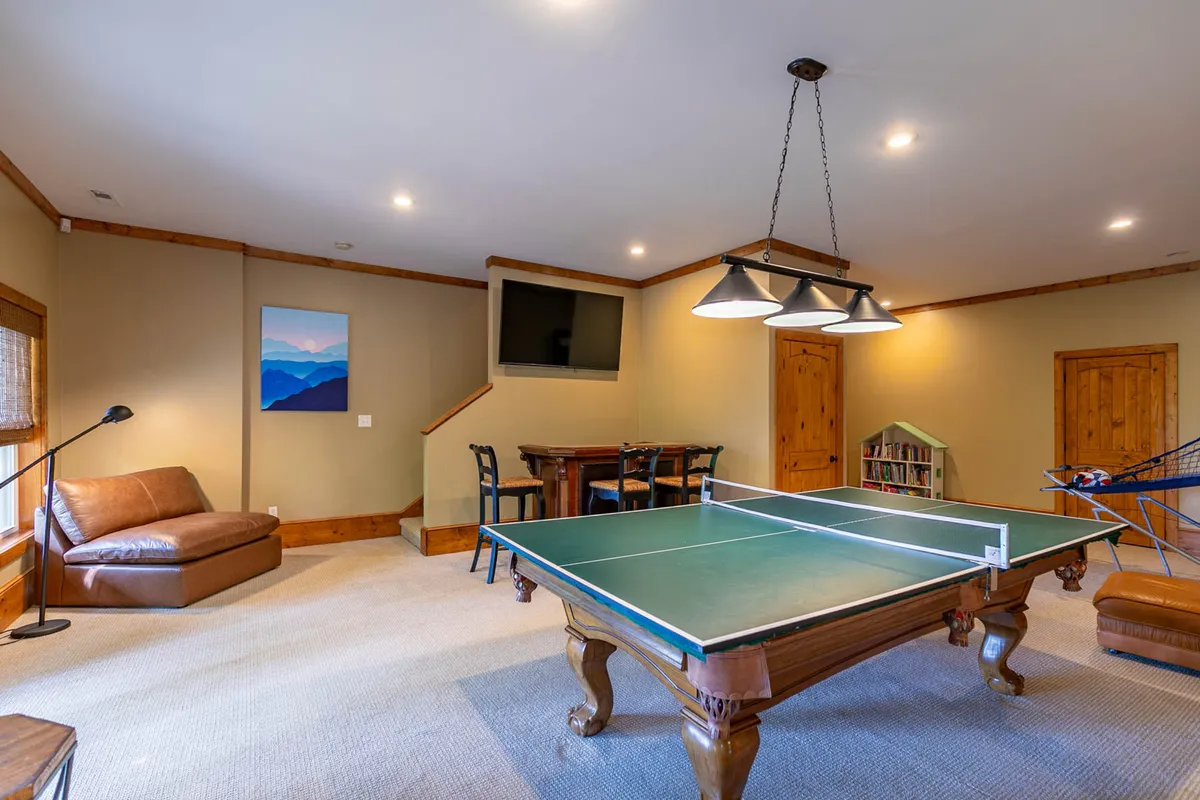 This lively game room scene features a centrally placed ping pong table, a comfortable leather couch for family and friends to relax, a wooden bar table perfect for board games or snacks, a wall-mounted TV for movie nights, and colorful artwork. The space is designed to encourage both casual hangouts and active play.