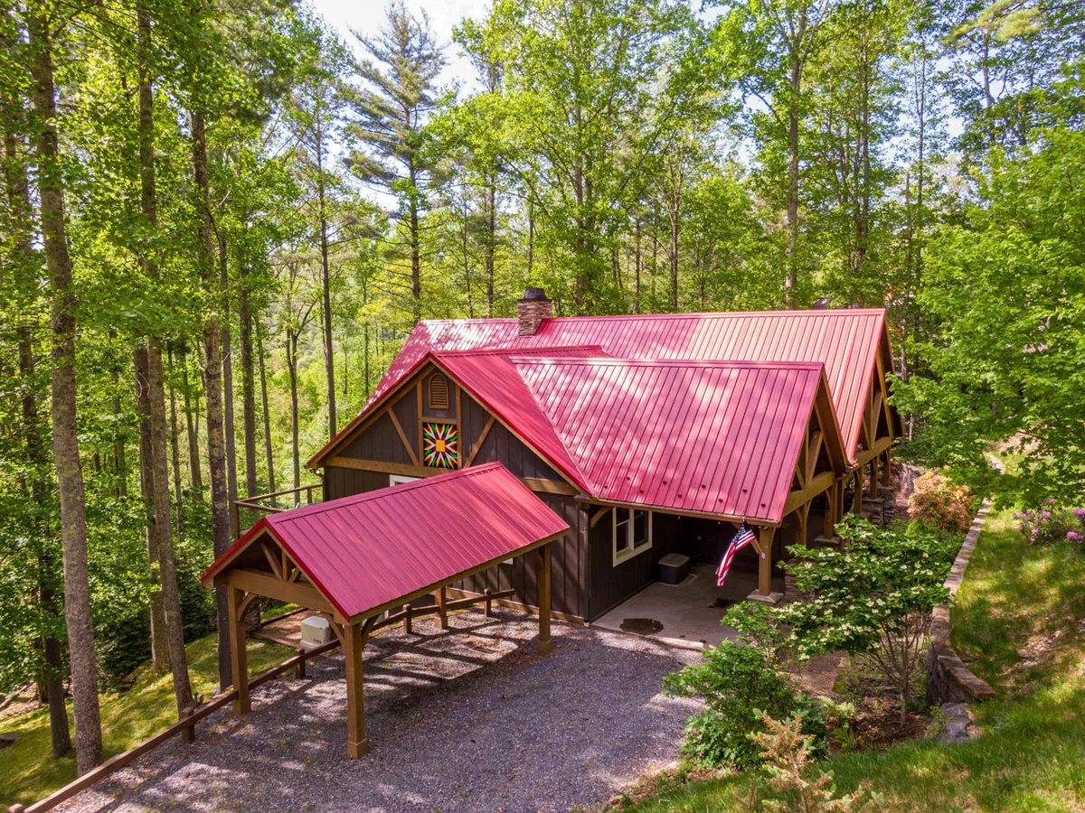 The image showcases the exterior of a spacious single-family mountain lodge in Boone, North Carolina, featuring a striking red metal roof, a welcoming wooden porch adorned with a colorful quilt square, and tranquil natural surroundings. The porch invites guests to relax, gather with loved ones, and enjoy the peaceful outdoor setting, with hints of nearby trails for exploration and open skies for stargazing.