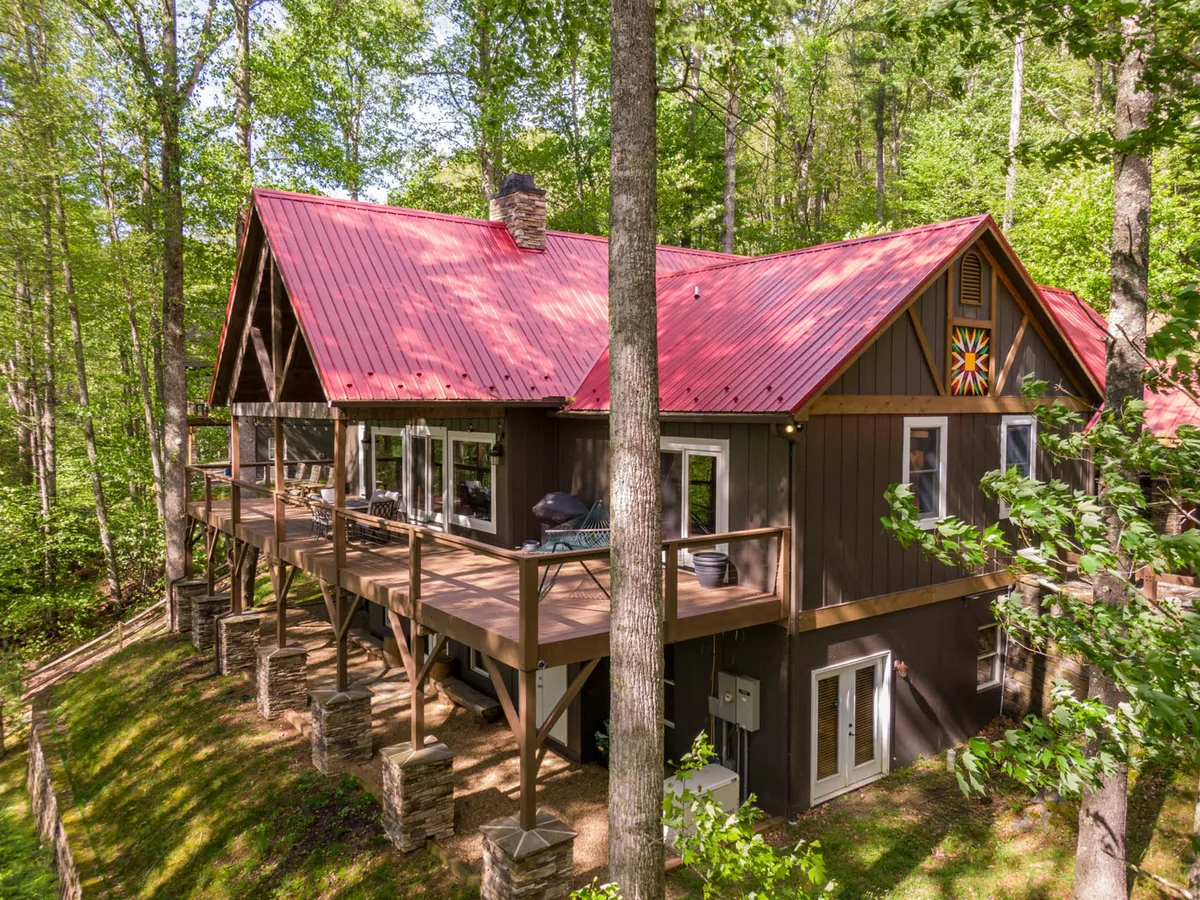 A spacious wooden deck with a red metal roof and sturdy stone pillars, overlooking wooded surroundings. Large windows connect the indoor and outdoor spaces, and the area invites guests to relax or gather after a day of activity.