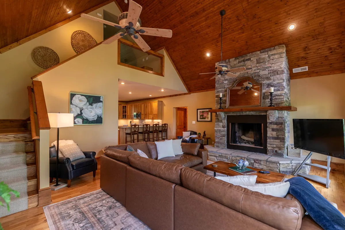 A seamless open-concept living area with a stone fireplace, spacious leather sofa, wood-accented ceiling, and direct sight lines into a well-appointed kitchen area. The arrangement encourages both relaxation and social interaction.